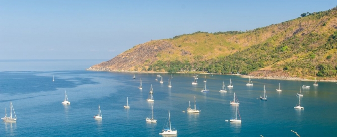 View of Nai Harn Beach, Phuket, with turquoise waters and numerous yachts moored offshore under a clear blue sky.